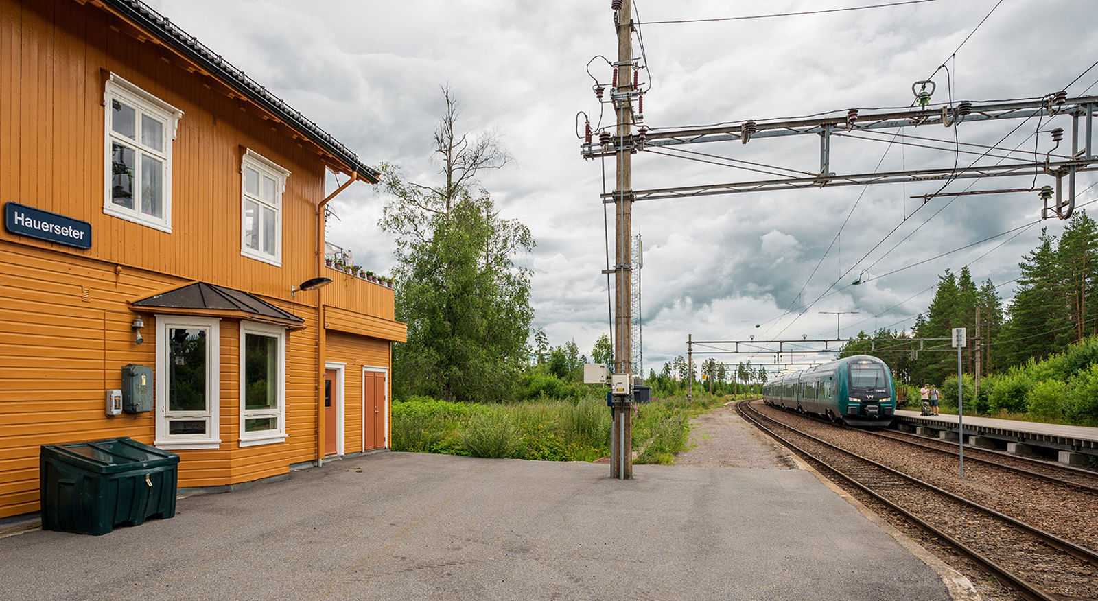 Gul stasjonsbygning, plattform og Vy-tog på sporet.