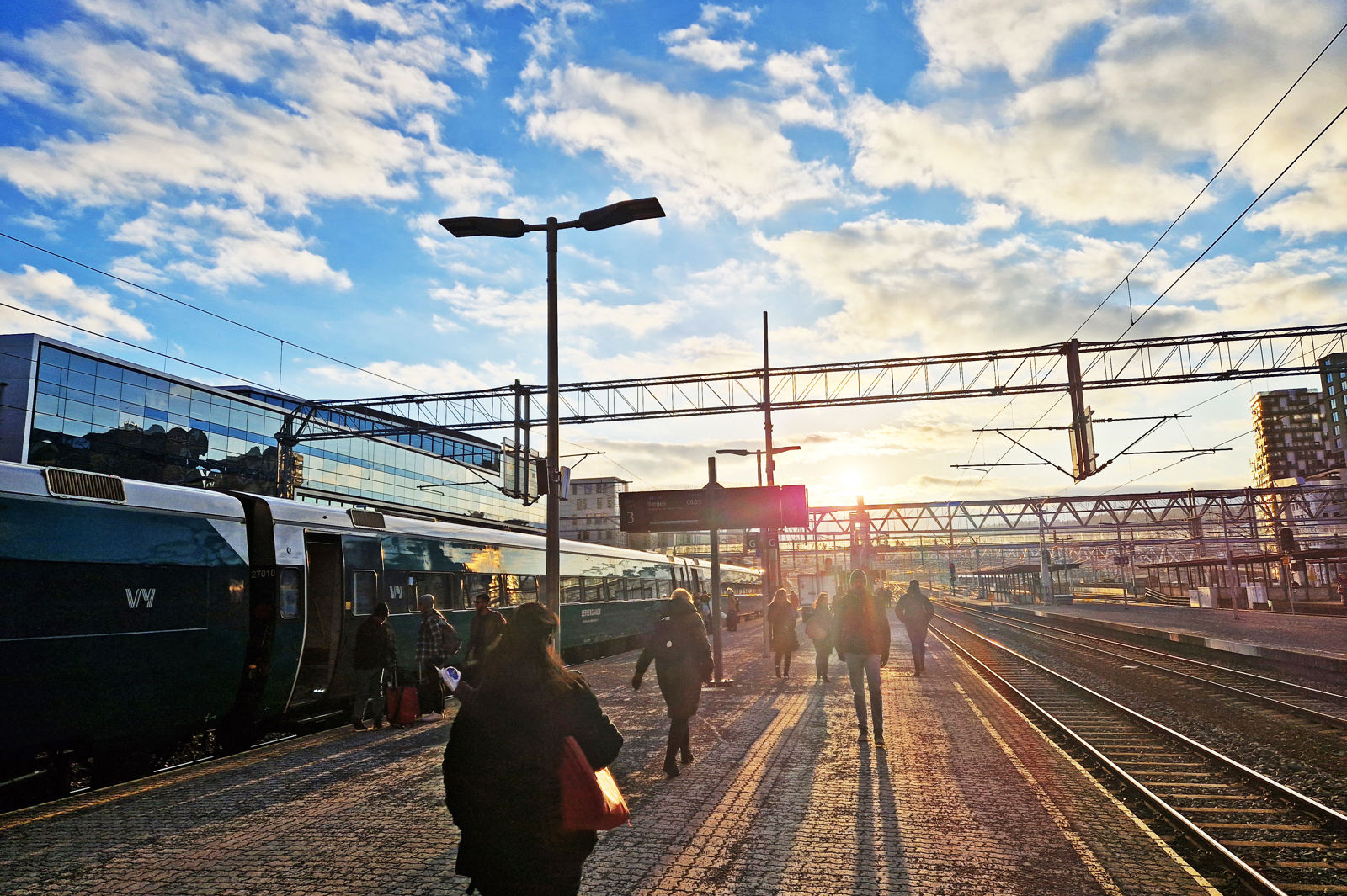 Lav sol over perrongene på Oslo S. Folk går på plattformen mot sola. Et tog står på ett av sporene.