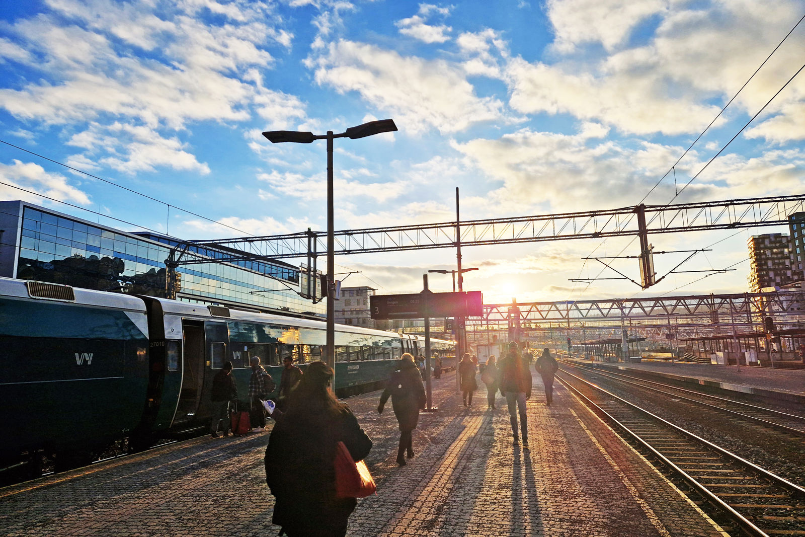 Lav sol over perrongene på Oslo S. Folk går på plattformen mot sola. Et tog står på ett av sporene.