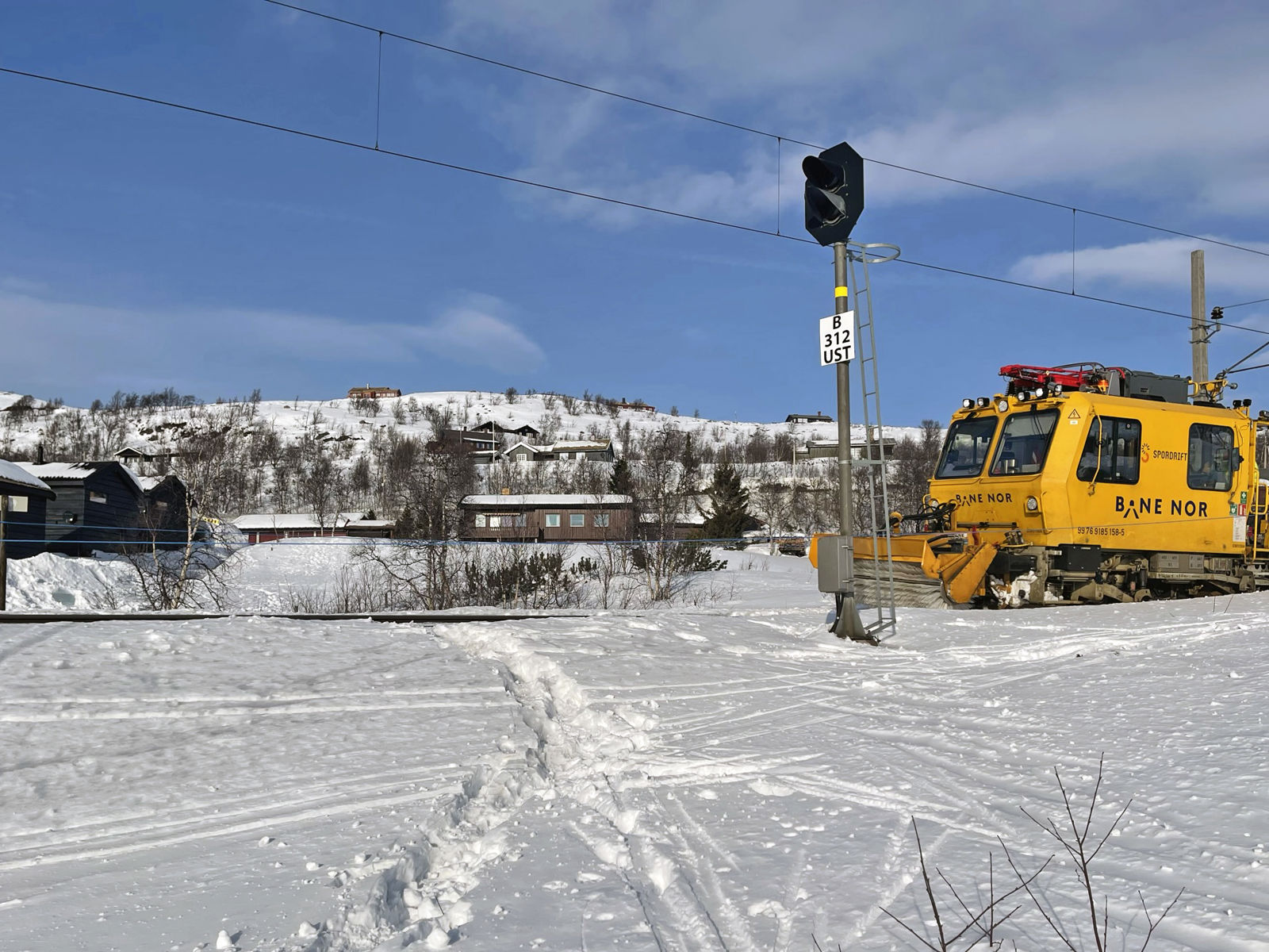 Spor i snøen krysser jernbaneskinnene. En gul arbeidsmaskin fra Bane NOR står på sporet.