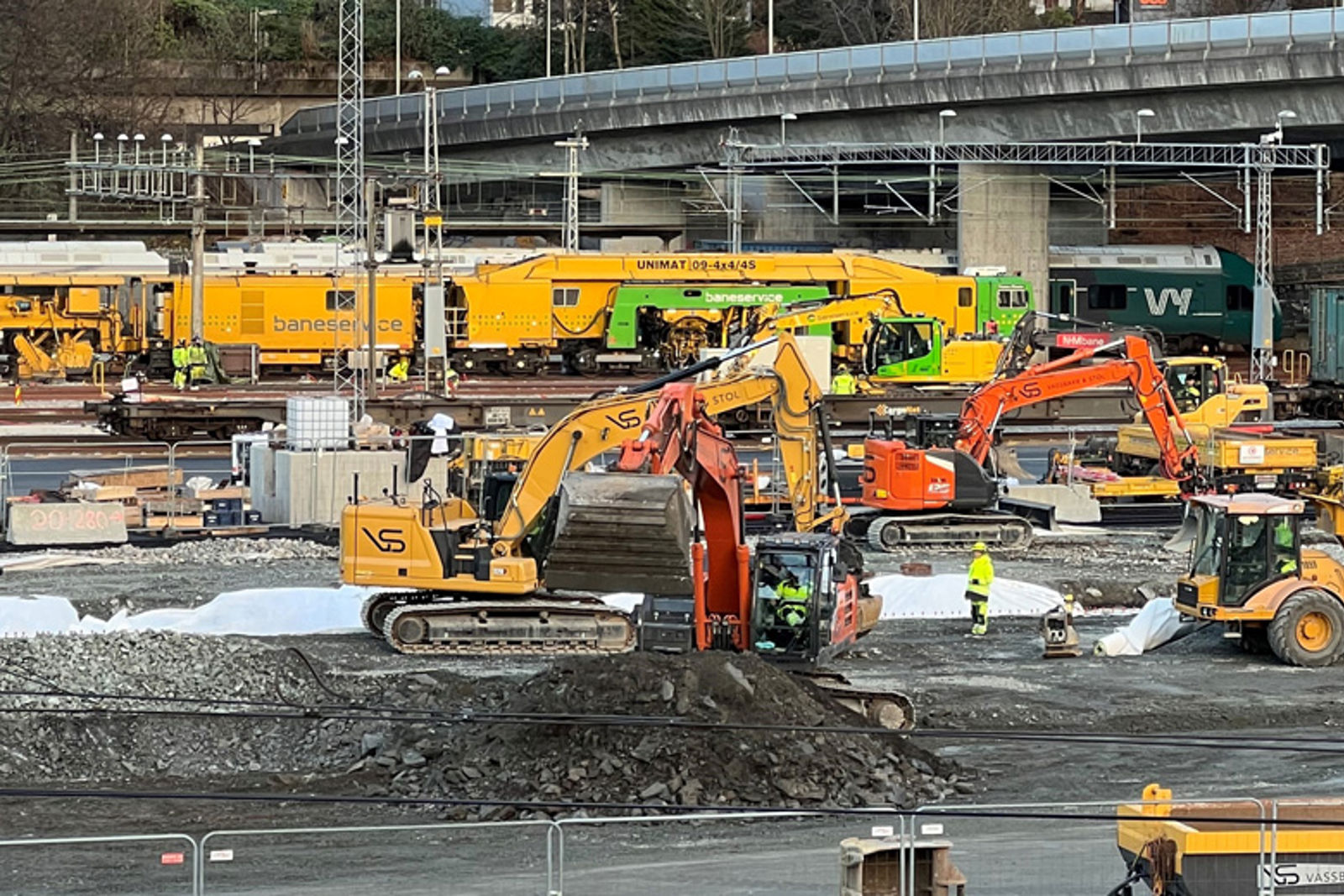 Arbeidsmaskiner inne på et jernbaneområde med tog i bakgrunnen