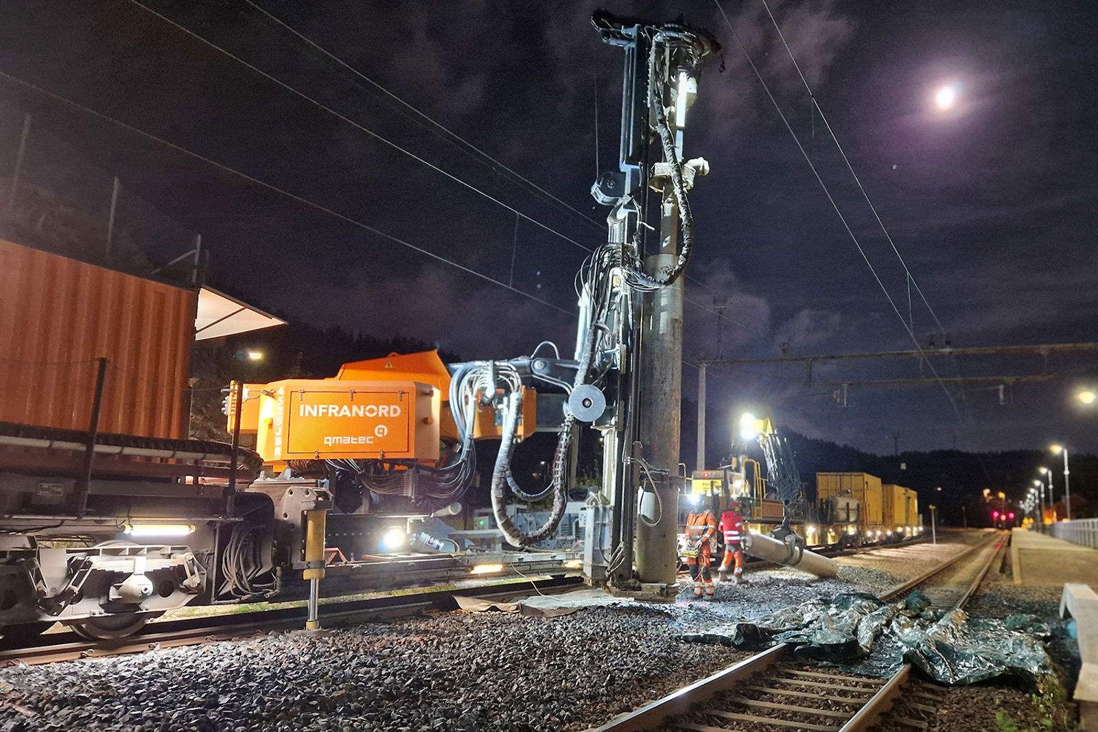Boring pågår mellom to spor på en stasjon med et stort spesialtog.  Det er natt og måneskinn.