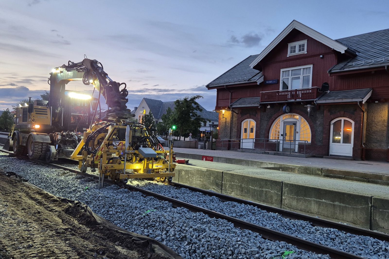 Gul gravemaskin jobber i jernbanespor foran rød stasjonsbygning.