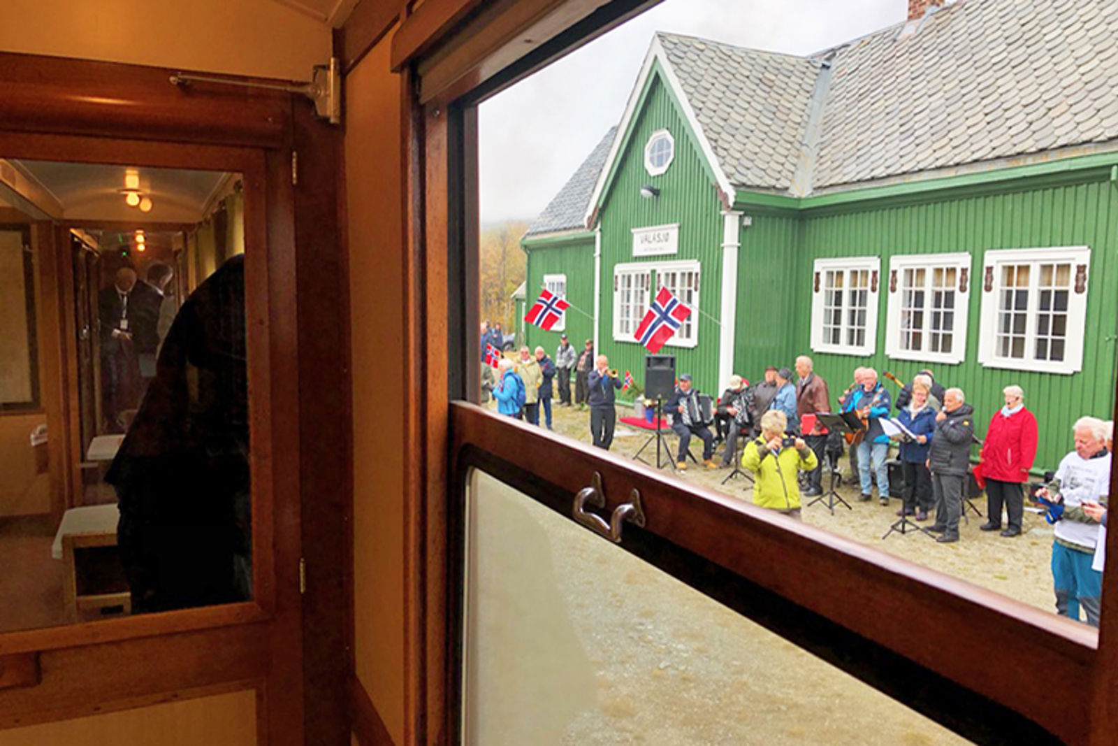 Bilde tatt innenfra et gammelt tog og ut, der vi ser en perrong med norske flagg og flere tilskuere.