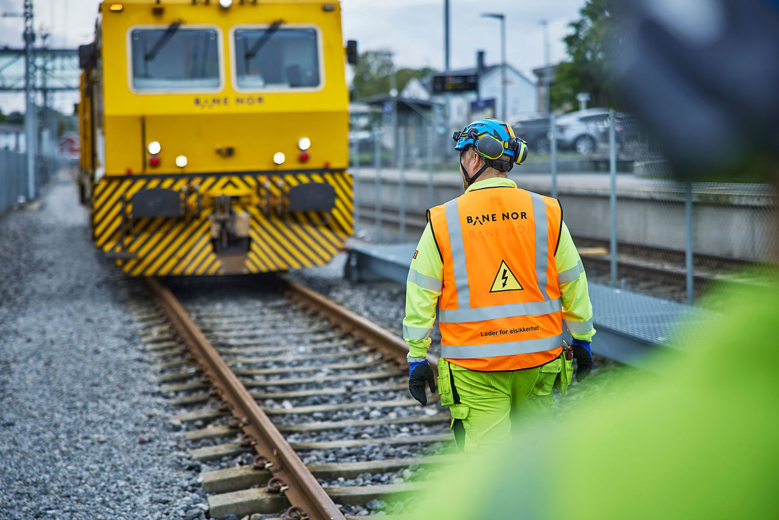 Leder for elsikkerhet med oransje vest i sporet, gult arbeidstog i bakgrunnen.