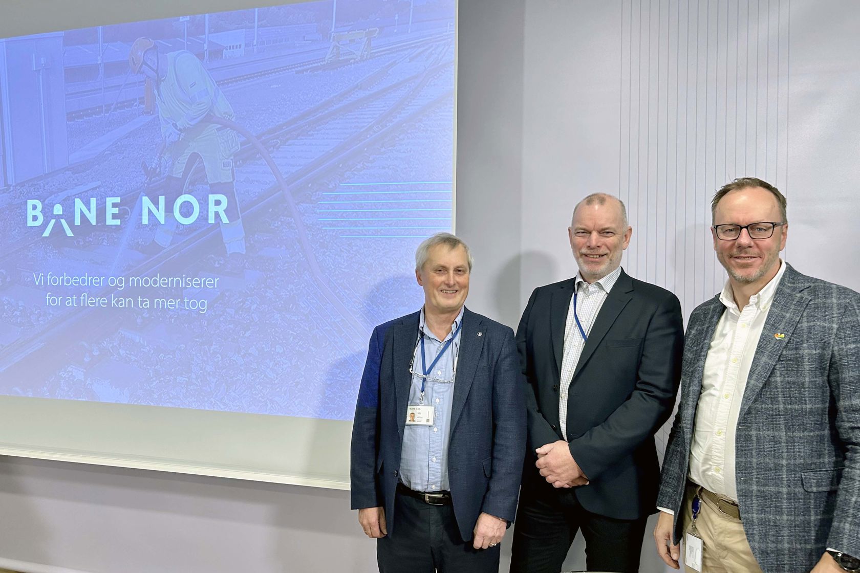 Tre menn i dress smiler fremfor skjerm som viser presentasjon